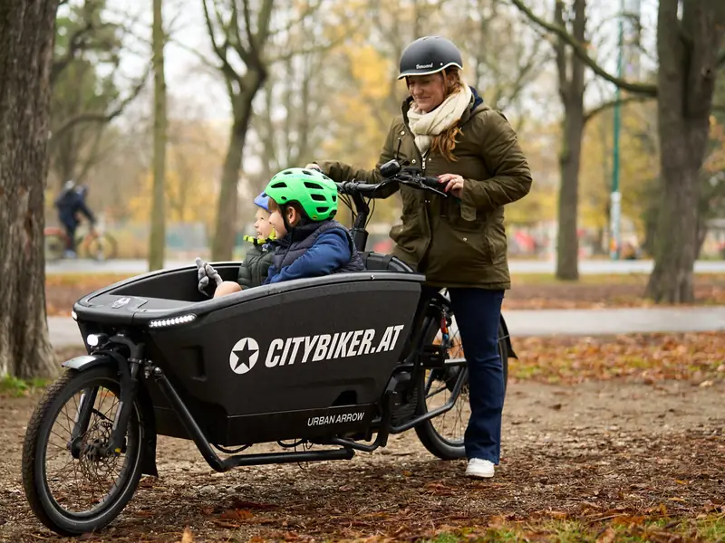 Une femme à côté de son vélo cargo dans un parc en automne, deux enfants assis à l'avant dans la caisse.